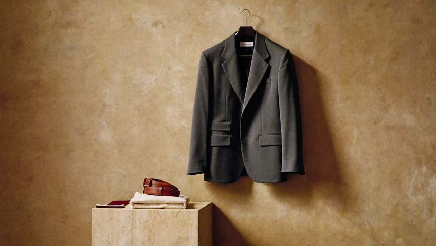 Un atelier con pareti di calce e travertino: tavolo in noce con maglie piegate e tre giacche su un’asta