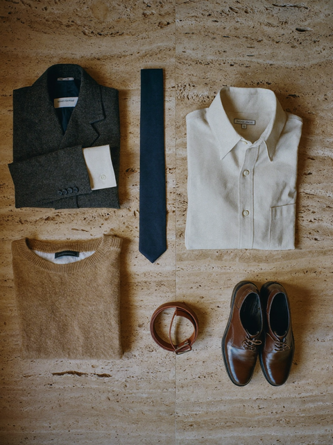 Composizione dall’alto su travertino: blazer antracite piegato, camicia avorio, maglione color cammello, cravatta blu notte, cintura e scarpe oxford in pelle marrone