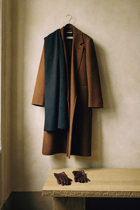 Lungo cappotto in lana color tabacco appeso a un piolo in noce su parete di calce, con sciarpa blu notte sulla spalla e guanti in pelle su una panca di travertino